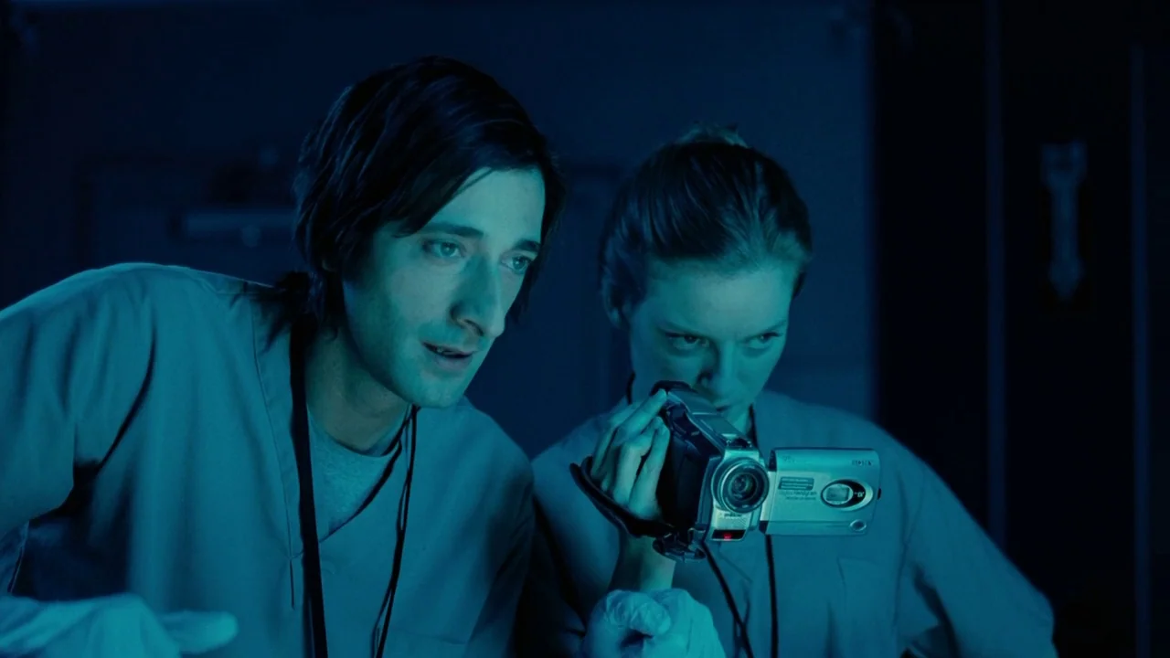 Splice (2009) movie still: medium — A dark, dimly lit environment with blue light casting deep shadows. A man and woman wearing scrubs look toward…