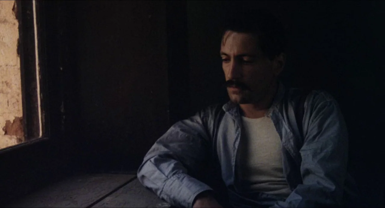 Breaker Morant (1980) movie still: medium — A dark, minimally lit room features a single window casting light from the left. A man sits by the…