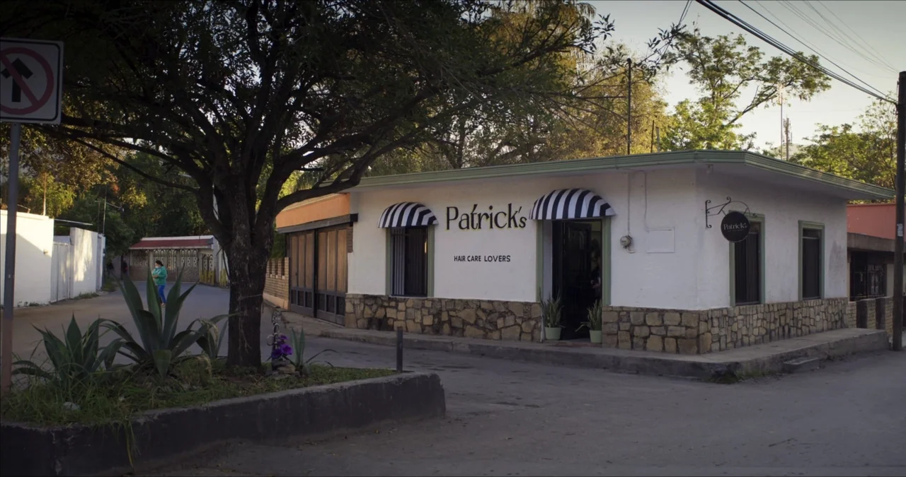 Bendita Suegra (2023) movie still: establishing — A single-story white hair salon building features stone-clad walls and black-and-white striped…