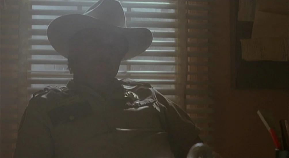 Near Dark (1987) movie still: medium — A man in a uniform and cowboy hat sits before a window with blinds. The bright light from outside puts the…