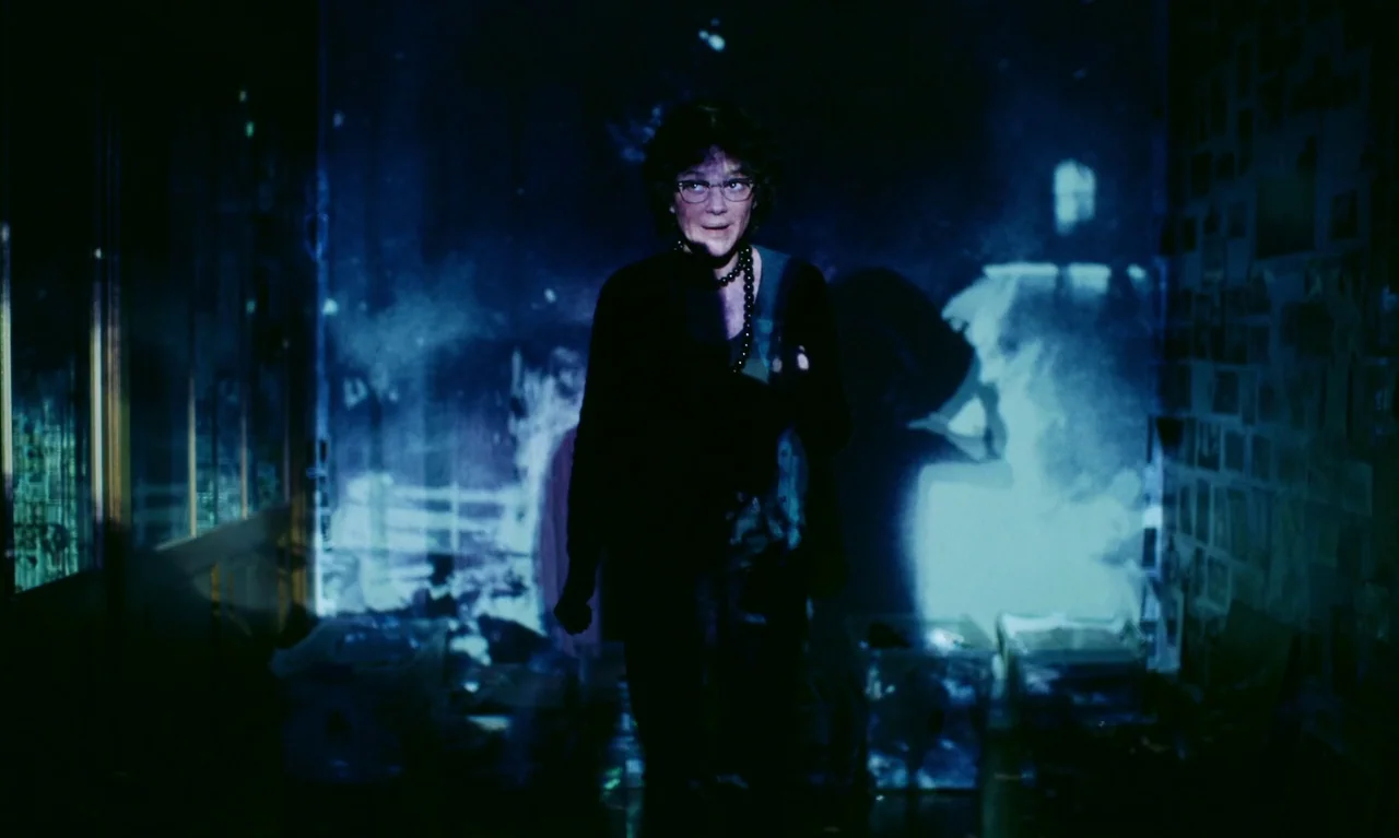 Little Girl Blue (2023) movie still: medium — A woman stands in a darkened room with blue projections casting light onto her face. Multiple images…