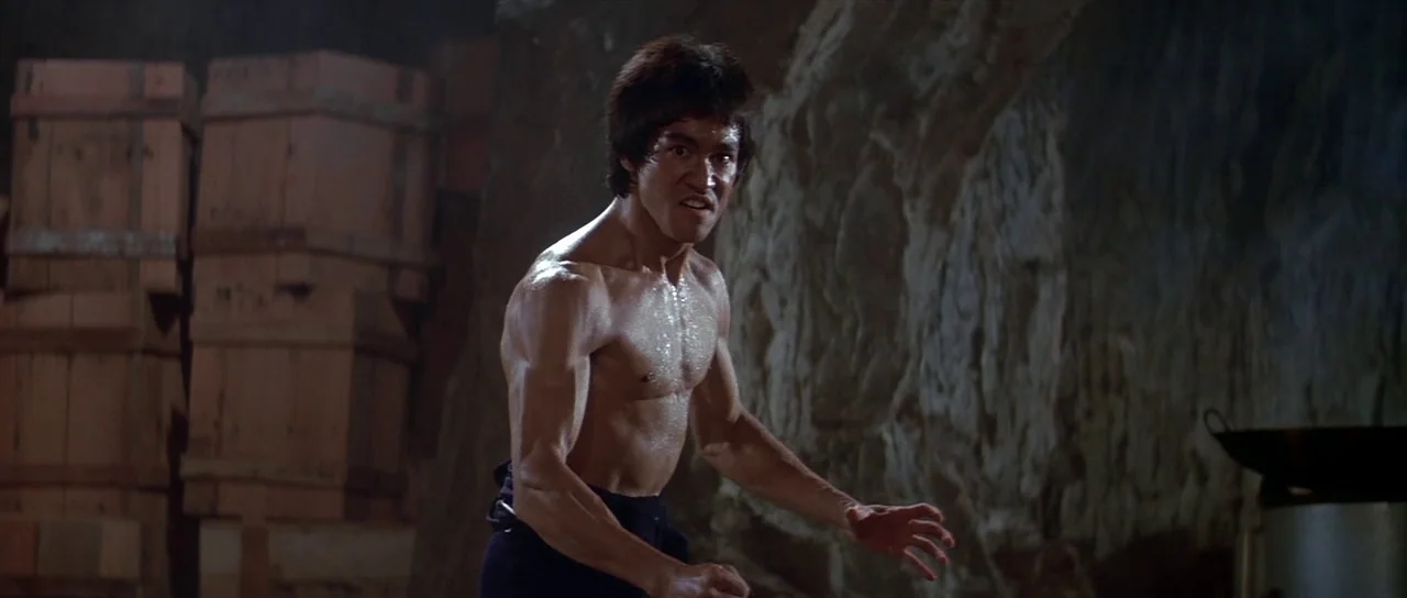 Enter The Dragon (1973) movie still: medium — A dimly lit rocky cavern interior contains large wooden shipping crates and stone walls. An athletic…
