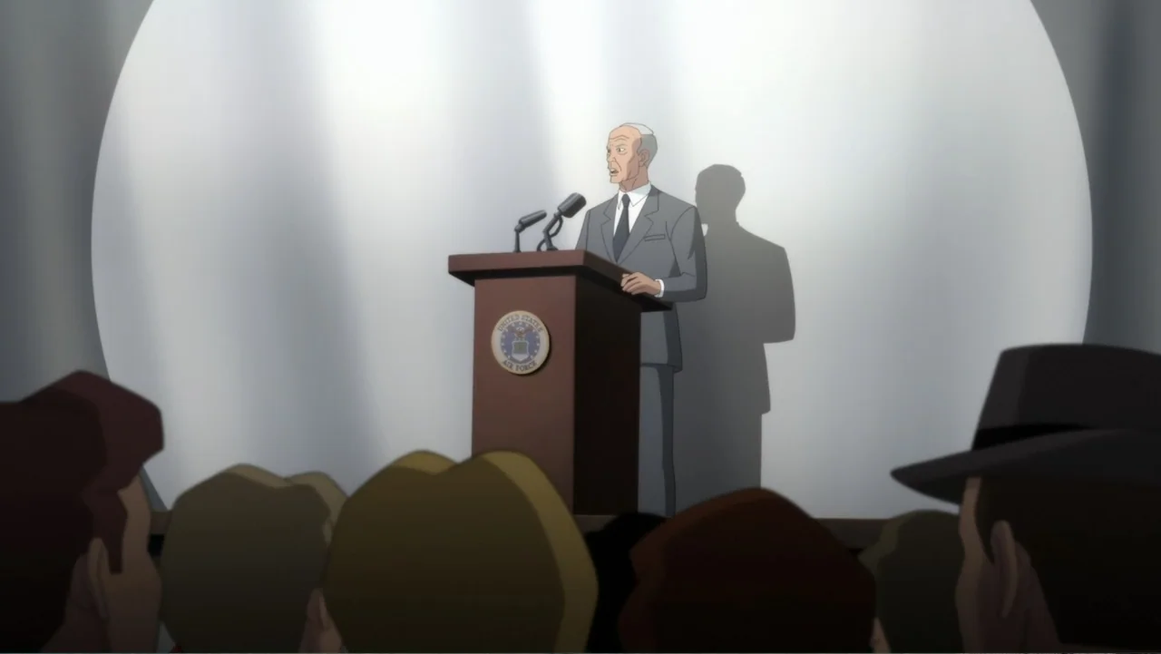 Superman: Red Son (2020) movie still: medium — An elderly man stands at a wooden podium in a stark, brightly lit space. He addresses an audience…