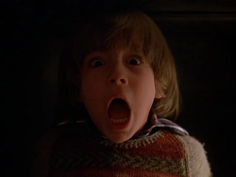 The Shining (1980) movie still: closeup — A child is positioned in deep shadow against an undefined dark backdrop. The boy displays a wide-eyed,…