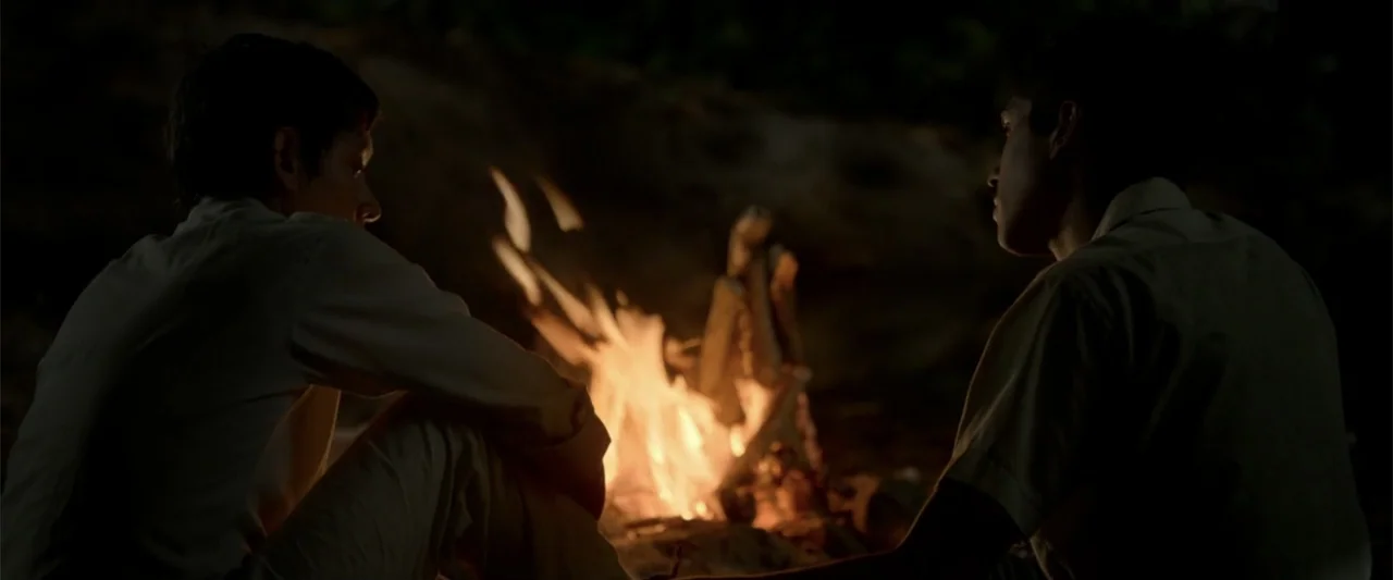 I Dream in Another Language (2017) movie still: medium — An outdoor campsite at night features a small fire burning in the center. Two men sit on…