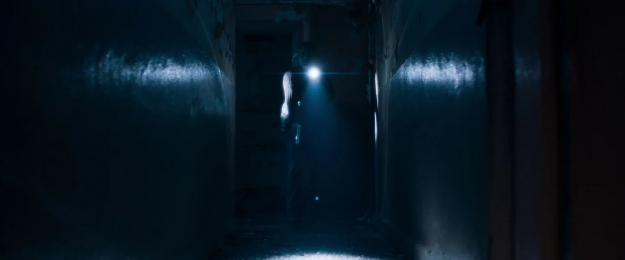 Baskin (2015) movie still: wide — A narrow, dark building corridor features dilapidated walls with peeling paint and shadows. A man holding a bright…