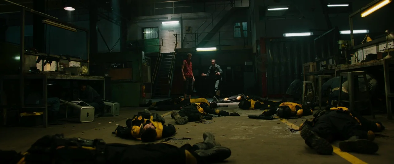 Boy Kills World (2023) movie still: wide — A dimly lit industrial workspace contains several bodies scattered across a concrete floor. Two standing…
