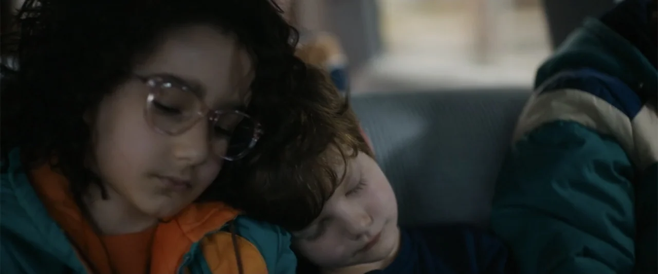 Unsung Hero (2024) movie still: closeup — A close view of a car backseat interior with soft natural lighting. Two children lean against each other…