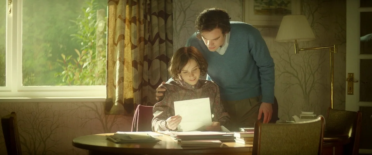 The Theory of Everything (2014) movie still: medium — A 1970s dining area features wallpaper with tree branch patterns and a wooden table. A man…