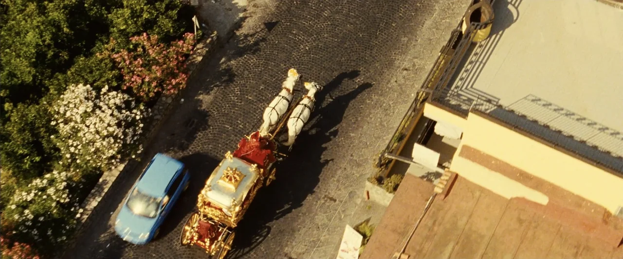 Reality (2023) movie still: wide — A top-down view shows a cobblestone road bordered by white flowering bushes and buildings. A horse-drawn gilded…