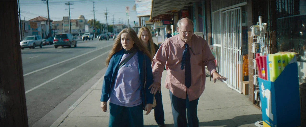 Kajillionaire (2020) movie still: fullbody — A suburban sidewalk runs alongside a busy street with parked cars and storefronts. Three people walk in…