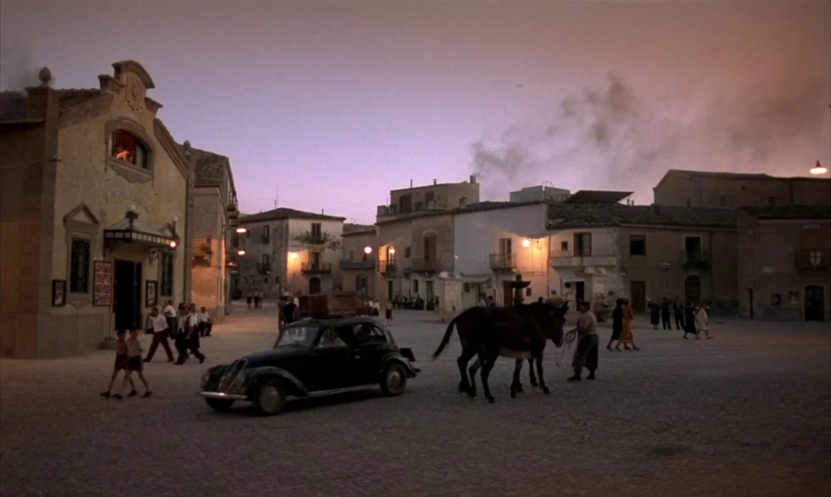 Cinema Paradiso (1988) movie still: wide — A quiet village square at dusk with stone buildings and a gravel surface. A small vintage car with…
