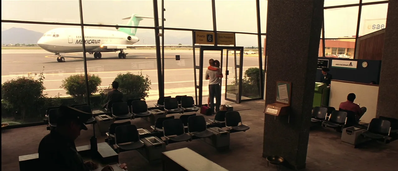 The Mexican (2001) movie still: wide — An airport boarding gate area features large windows overlooking an aircraft on the tarmac. A couple embraces…