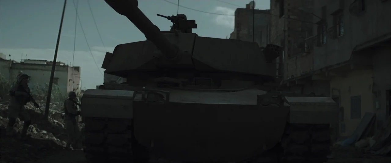 American Sniper (2014) movie still: medium — An M1 Abrams tank occupies the center of a narrow, rubble-strewn urban street. Soldiers stand in the…