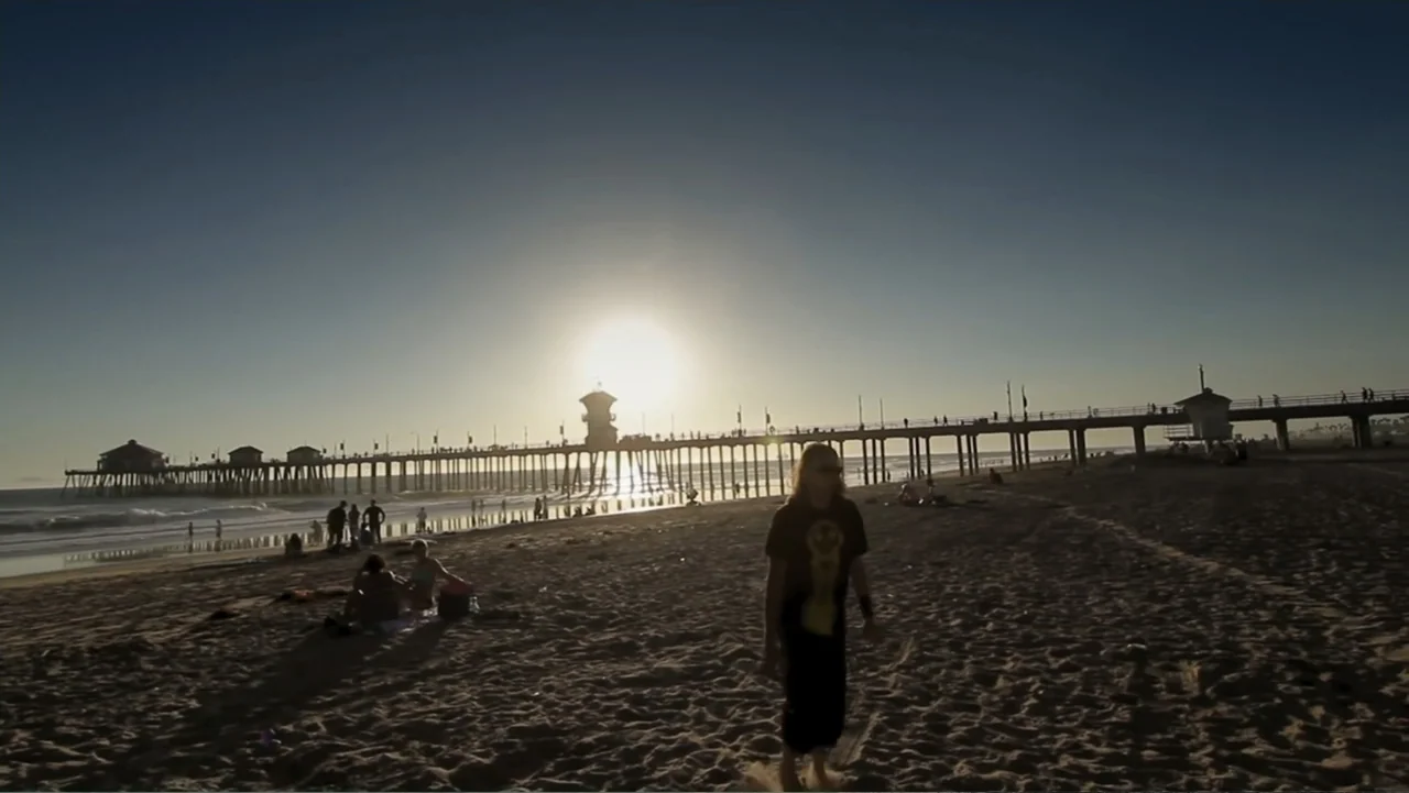 Control (2022) movie still: fullbody — A wide sandy beach stretches toward a long pier silhouetted against a setting sun. A person with long hair…