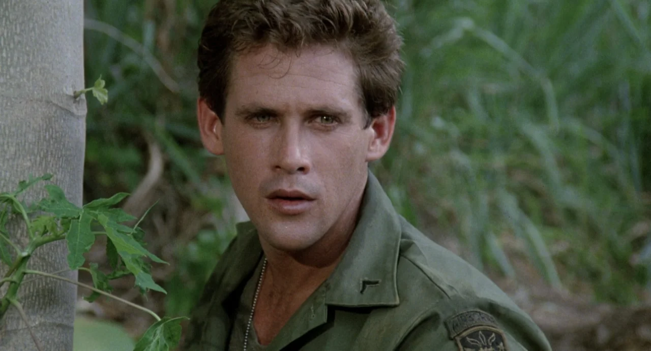 American Ninja (1985) movie still: closeup — A dense green jungle environment features a soldier positioned partially behind a tree trunk. The…