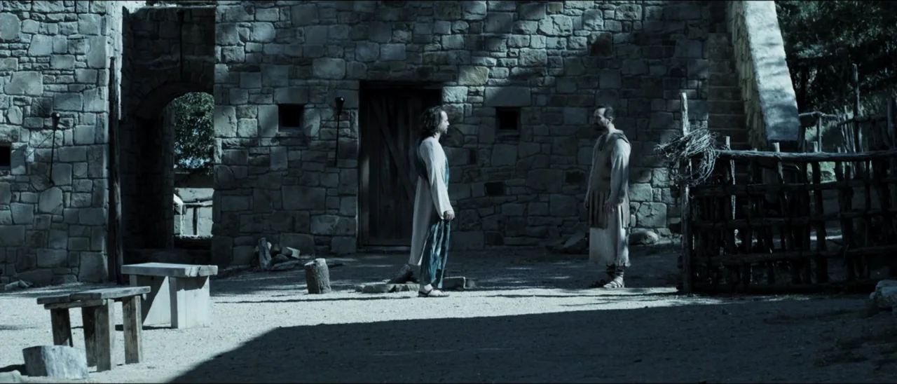 The Penitent Thief (2021) movie still: fullbody — A stone courtyard with a high wall and a large wooden door. Two men in ancient garments stand…