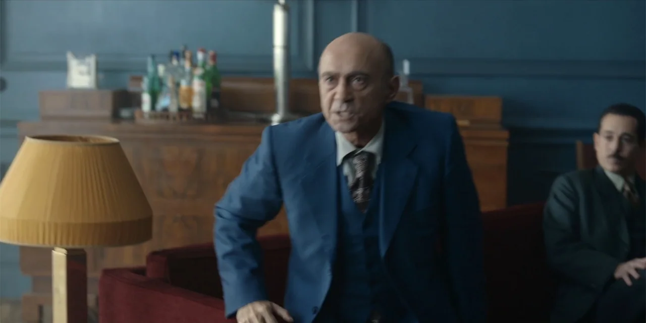 Oslo (2021) movie still: medium — A furnished interior room features dark blue walls and a red velvet sofa. A bald man in a suit stands talking to a…