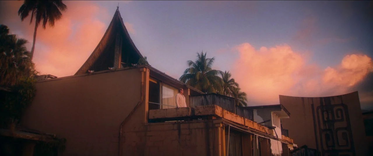 Pacifiction (2022) movie still: wide — A modern building with a steeply pitched, pointed roof stands against a sunset sky. A solitary man in a suit…