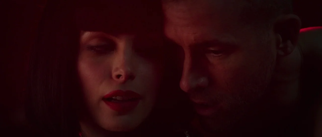 Deadpool (2016) movie still: closeup — A dimly lit interior space is saturated in intense red light. A woman with a dark bob and a man lean together…