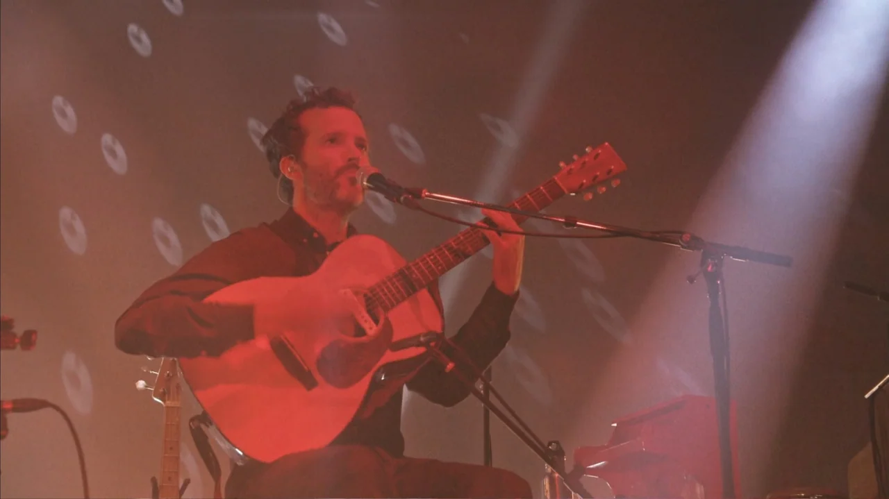 Flight of the Conchords: Live in London (2018) movie still: medium — A man performs on a dark concert stage under a focused spotlight. He sits while…