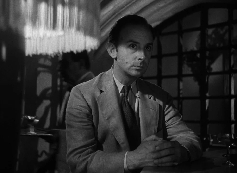 Casablanca (1942) movie still: medium — A dimly lit restaurant setting features a paneled window and a decorative hanging lamp. A man sits at a…