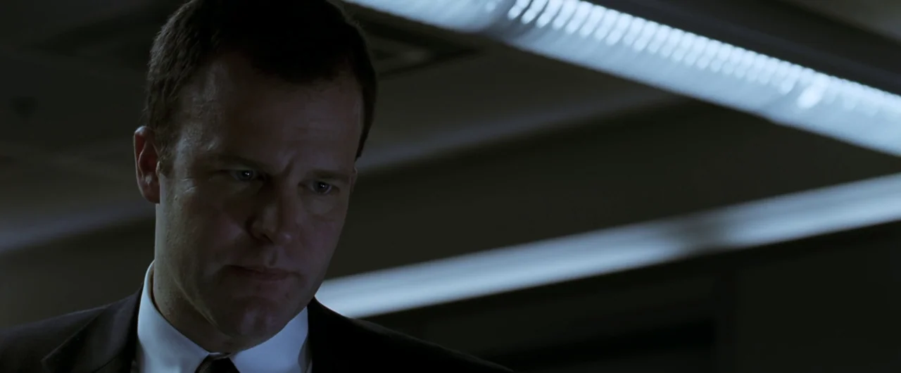 Syriana (2005) movie still: closeup — An interior space with a dimly lit, dark background featuring horizontal overhead fluorescent light. A man in…