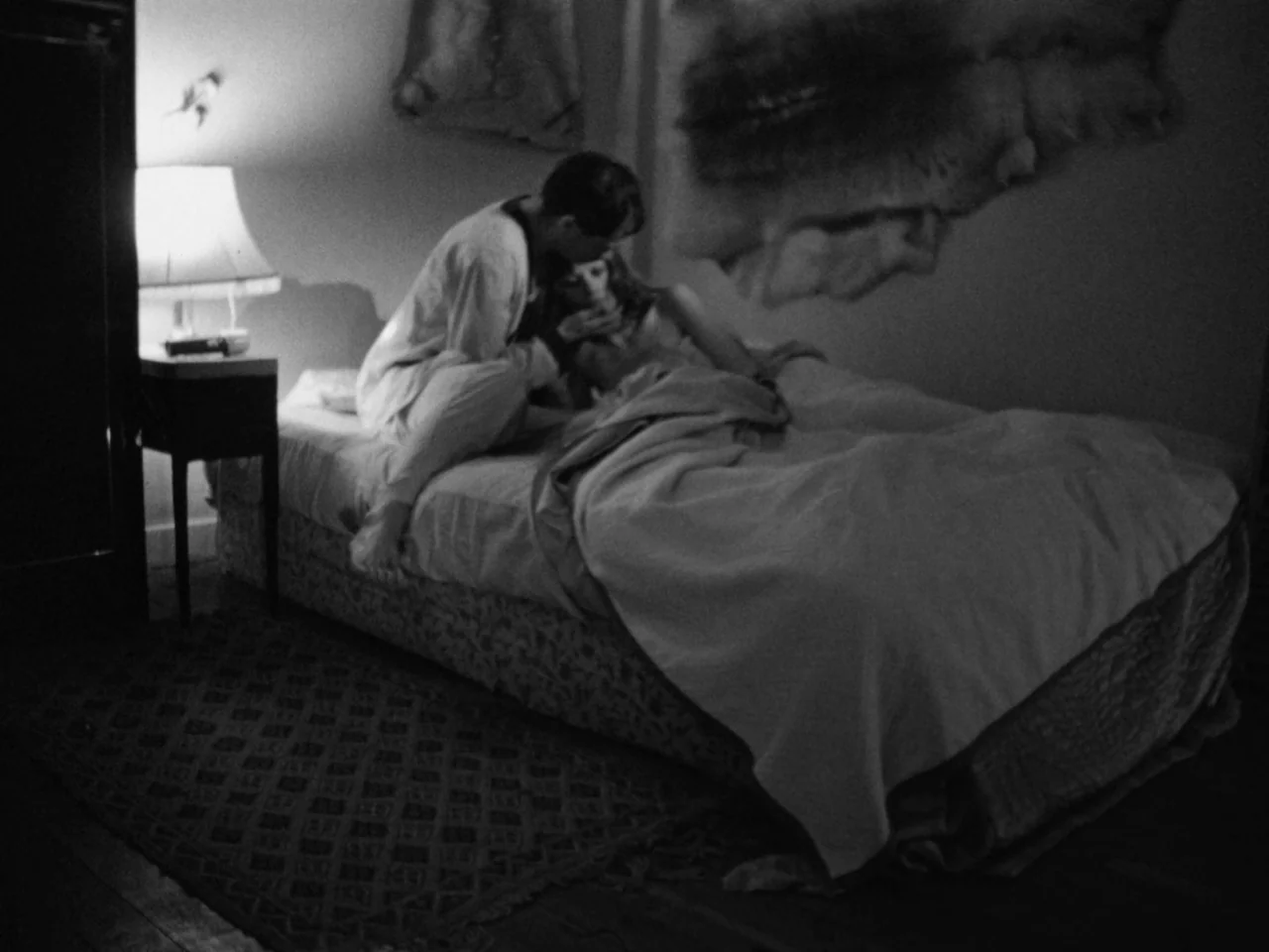 A Woman Kills (1968) movie still: medium — A dim room contains a bed with a person lying down under a blanket. A man sits on the edge of the…