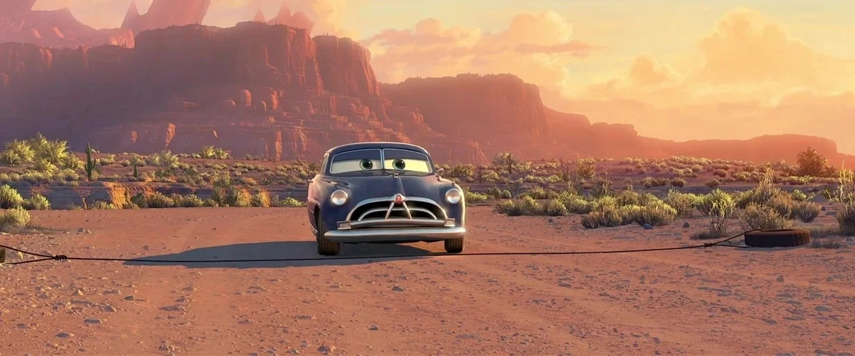 Cars (2006) movie still: wide — An arid desert landscape features red mesas under a golden late afternoon sky. An anthropomorphic vintage car pulls…