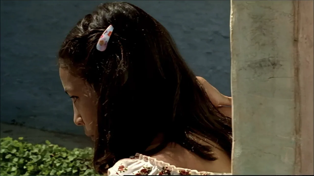 In the Middle of the World (1990) movie still: closeup — A woman stands behind a light-colored door frame against a dark blue wall. She looks toward…