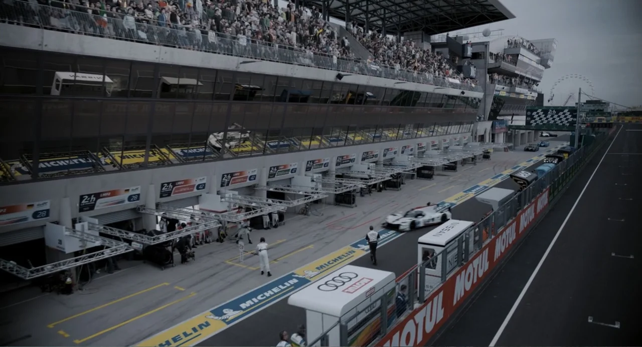Gran Turismo (2023) movie still: wide — A bustling racing pit lane features multiple garage bays and a large grandstand above. A white sports car…