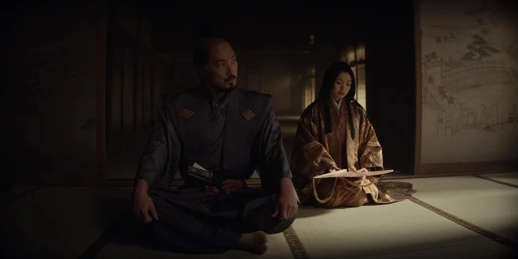 Shogun (2024) movie still: fullbody — A dark, spare room with tatami flooring and paper walls painted with landscape imagery. A man and woman sit…