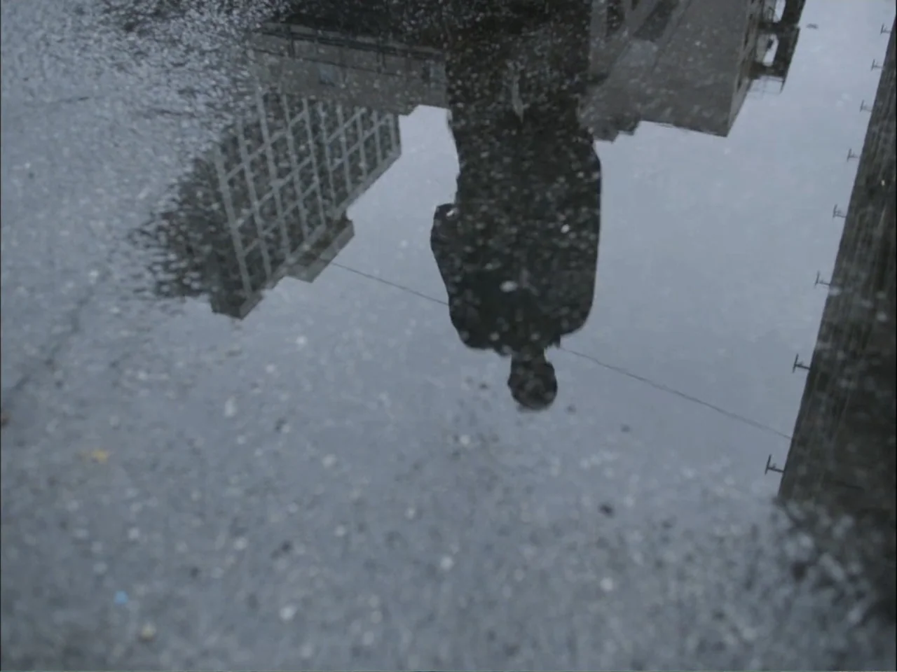 As We Speak: Rap Music on Trial (2024) movie still: wide — A rain-filled puddle on gray pavement reflects the silhouette of a person and buildings.…