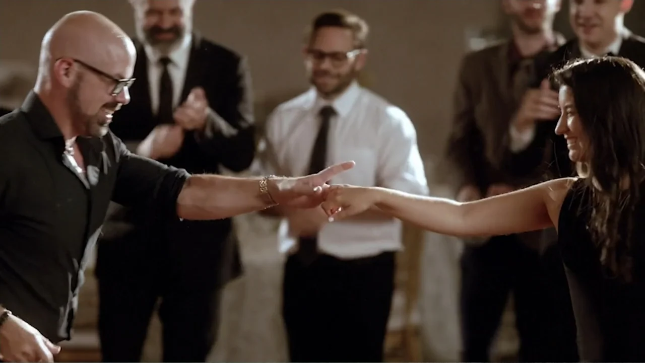 Ask Me to Dance (2022) movie still: medium — A group of people gathers in a large indoor event space during a party. A bald man and a woman in a…