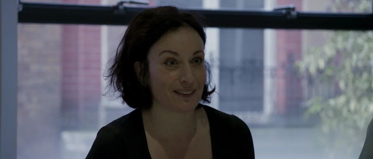 The Face of an Angel (2014) movie still: closeup — A woman stands in a bright indoor space with a large window behind her. She smiles while looking…