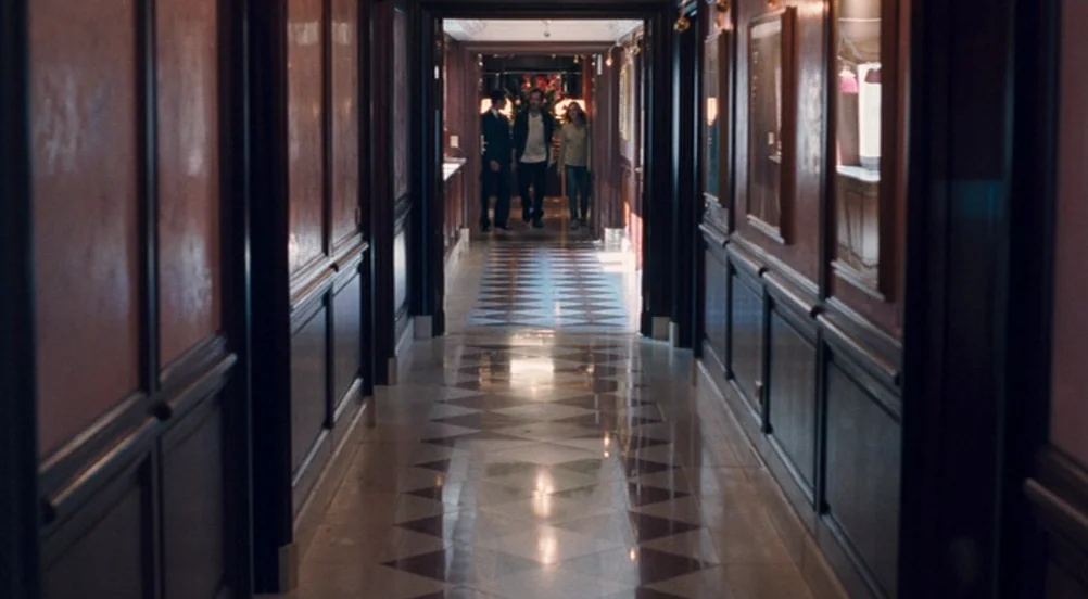 Somewhere (2010) movie still: wide — A long, narrow hotel corridor features dark wood paneling and a checkered marble floor. Three people walk in a…