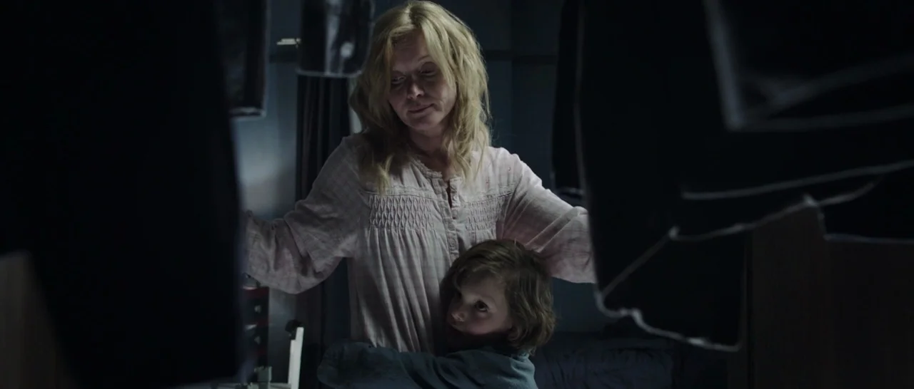 The Babadook (2014) movie still: medium — A dimly lit interior space filled with hanging clothes obscuring parts of the room. A woman in a nightgown…