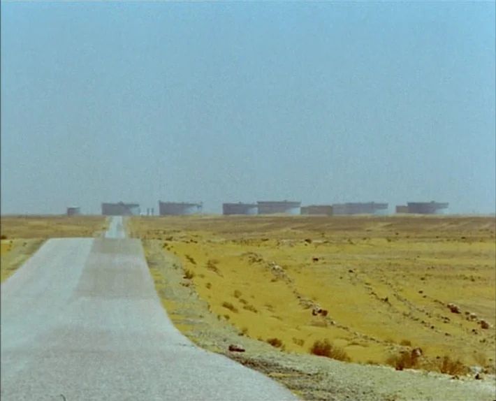 Fata Morgana (1971) movie still: wide — An asphalt road stretches into a flat, arid landscape under a clear sky. A line of cylindrical oil storage…