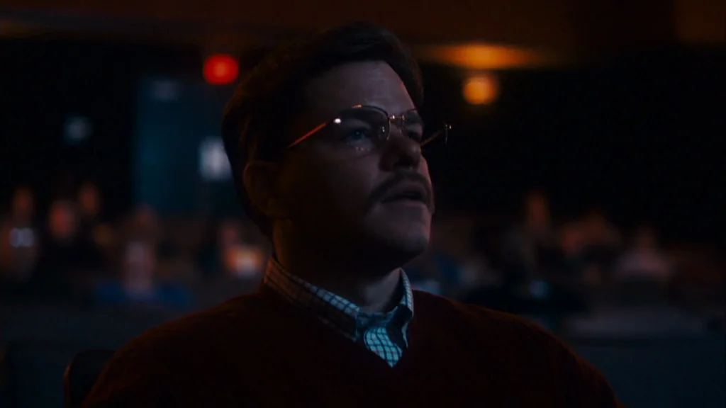 The Informant! (2009) movie still: medium — A man sits in the dim rows of a dark movie theater auditorium. He is watching a film while seated in his…