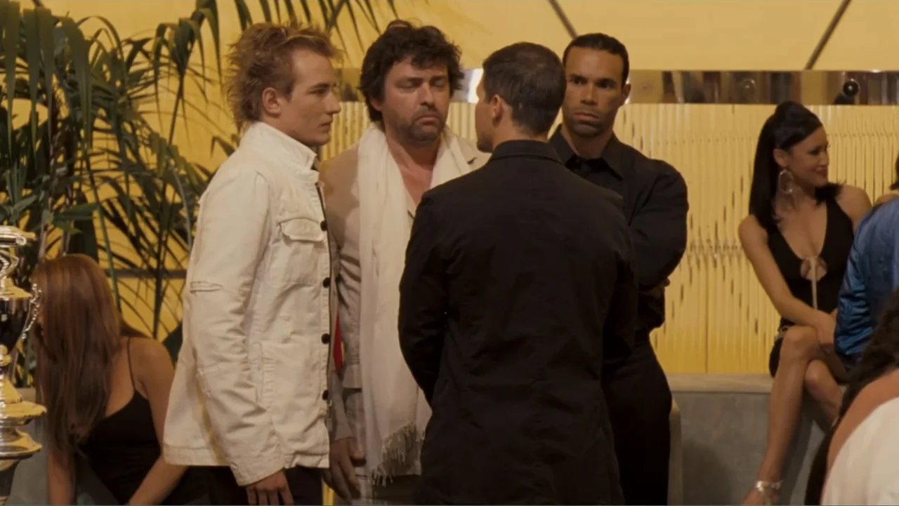 Redline (2007) movie still: medium — A crowded social venue with gold walls and potted palm plants at night. Several individuals stand near each…