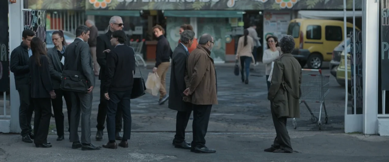 Doublespeak (2021) movie still: wide — A paved supermarket parking lot shows a group of business professionals standing together. Several adults in…