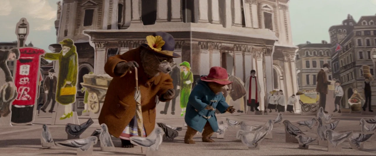 Paddington 2 (2017) movie still: wide — A miniature world constructed from paper cutouts replicates a bustling European city plaza. Two…