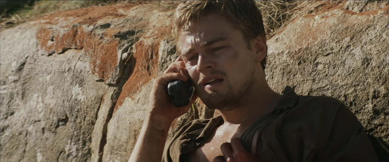 Blood Diamond (2006) movie still: medium — A man leans against a sun-drenched, textured rock face outdoors. He holds a black cell phone to his ear…