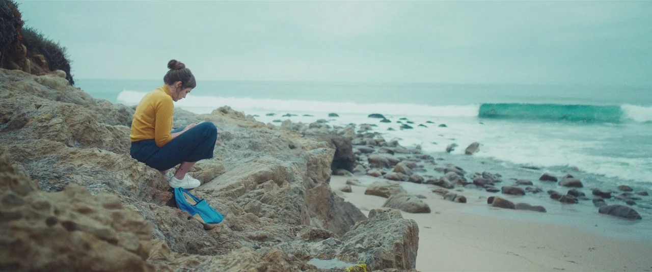 Gemini (2017) movie still: wide — A woman sits on jagged coastal rocks beside an overcast sea with crashing waves. She is dressed in a yellow top…