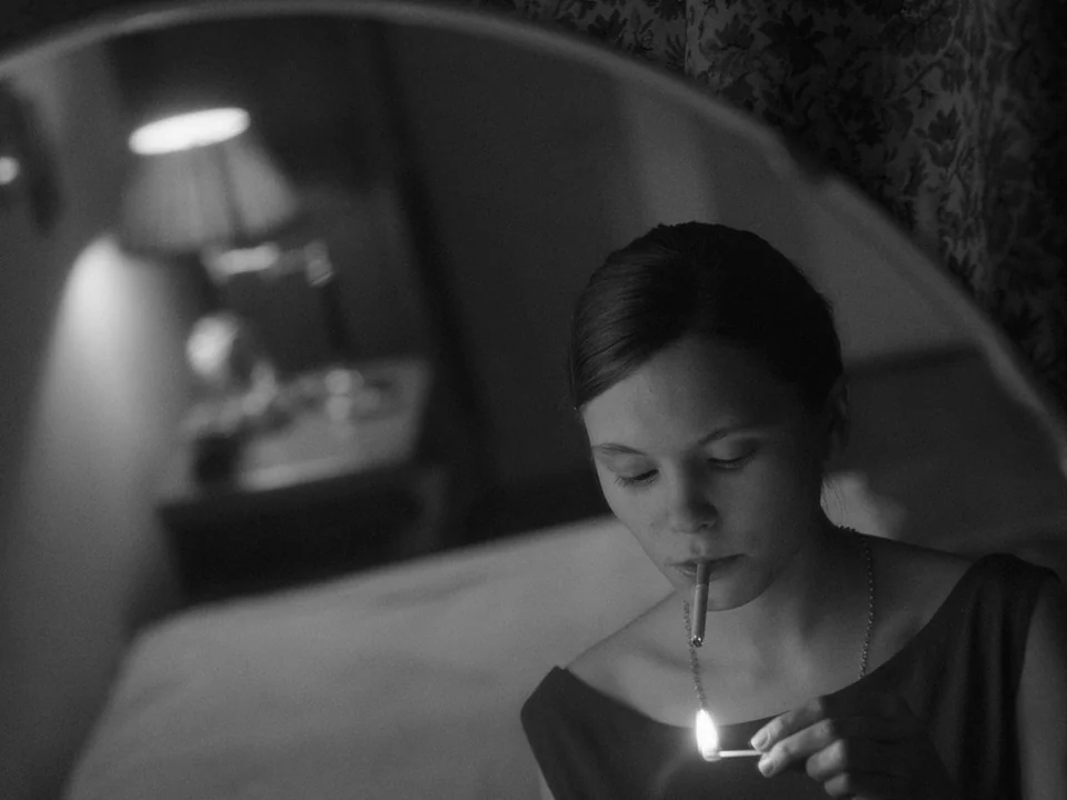 Ida (2013) movie still: closeup — A dimly lit room with vintage wallpaper visible in the mirror reflection. A woman uses a small wooden match to…