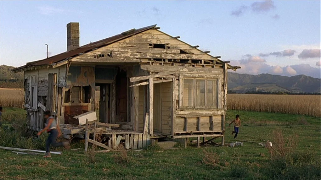 Boy (2010) movie still: wide — A dilapidated, weathered wooden house with peeling paint sits in a large open field. Two young boys run across the…