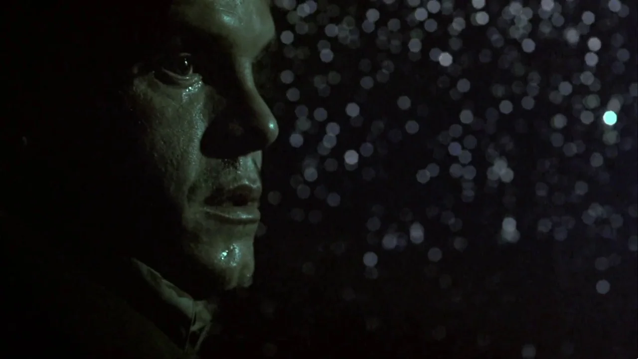 Hellraiser: Inferno (2000) movie still: closeup — A man stands in a dark urban environment during a night rain shower. Distant city lights create a…