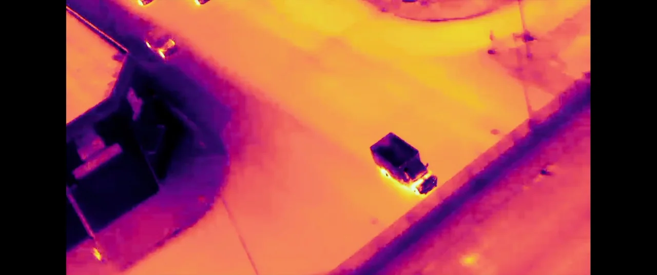 Ambulance (2022) movie still: wide — A thermal view from above shows a road and a corner of a building. A small boxy vehicle travels along the…