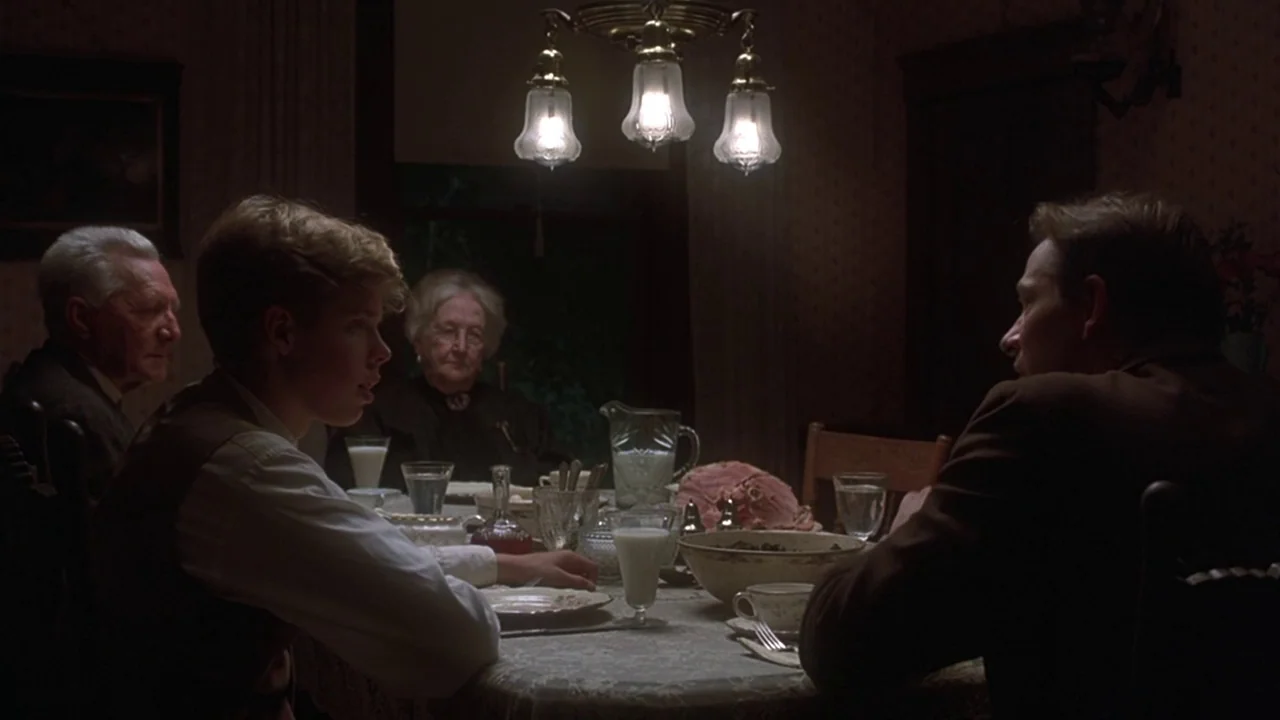 Matewan (1987) movie still: medium — A dark dining room with patterned wallpaper and a central multi-bulb chandelier. Four people are seated at a…