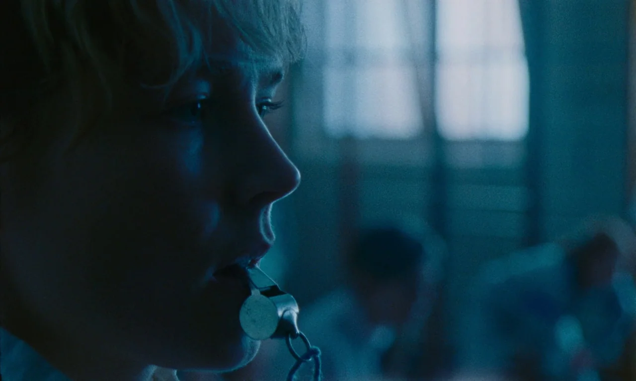 Blue Jean (2022) movie still: closeup — A dimly lit indoor gymnasium features a blurry background with window frames and other people. A blonde…